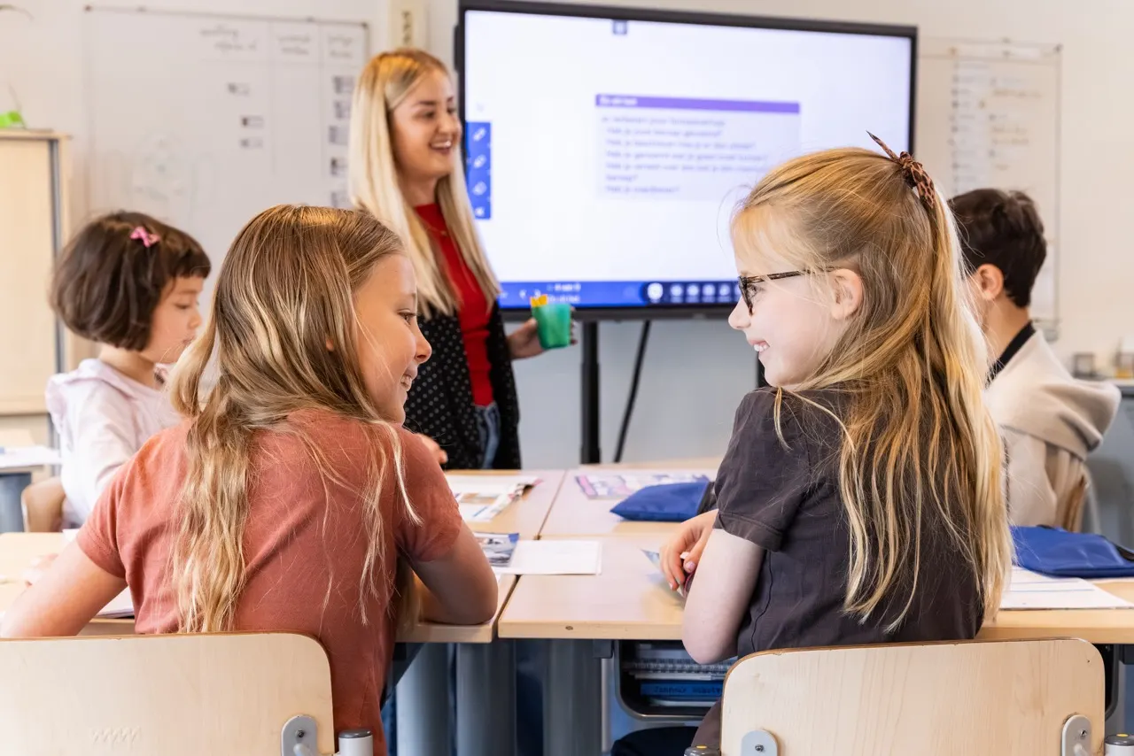 Twee leerlingen blij in de klas met leerkracht bij het digi bord